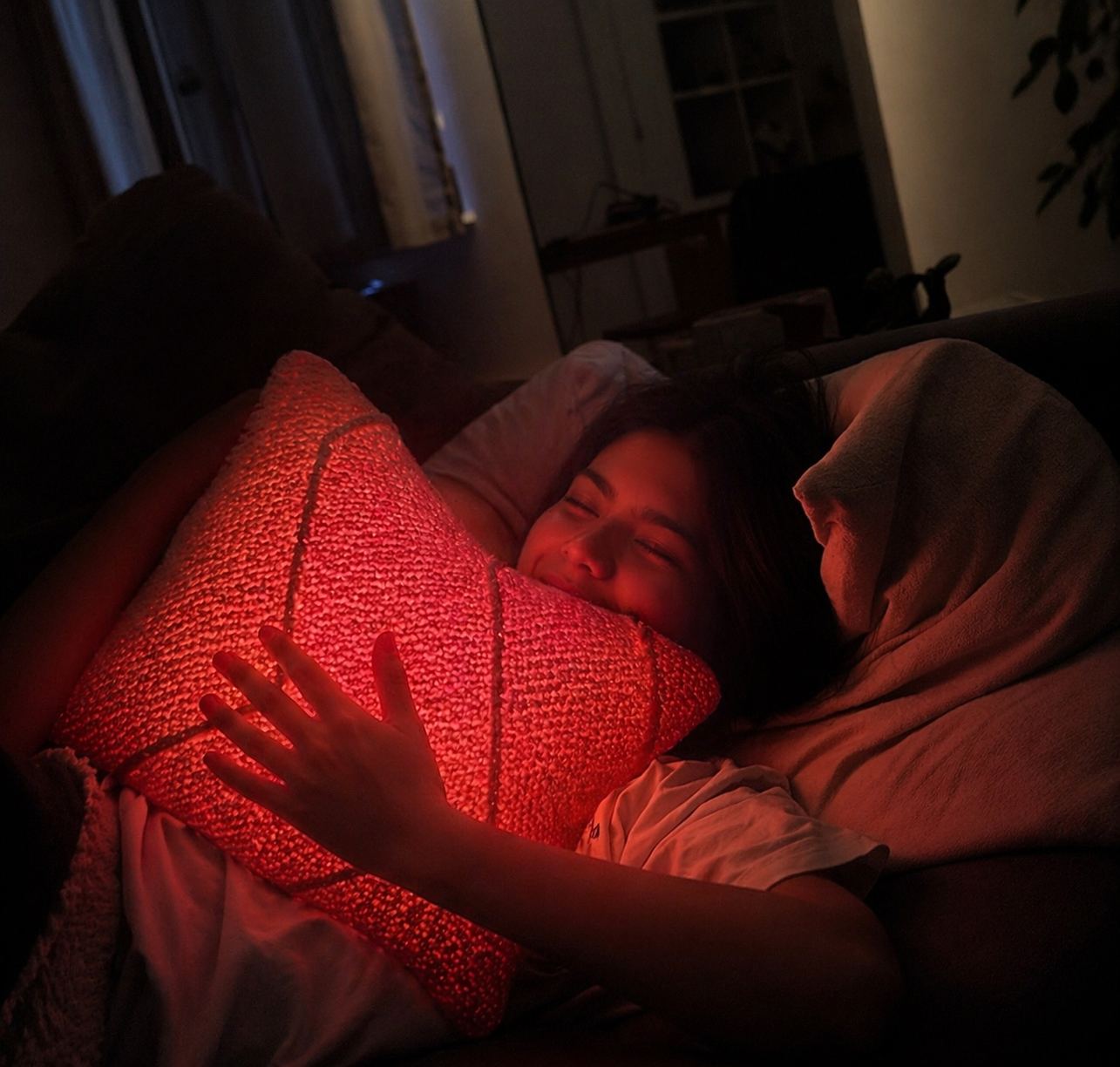 Two people apart, connected by light — a person on a sofa holding a Held by Miru cushion that glows warm from inside.